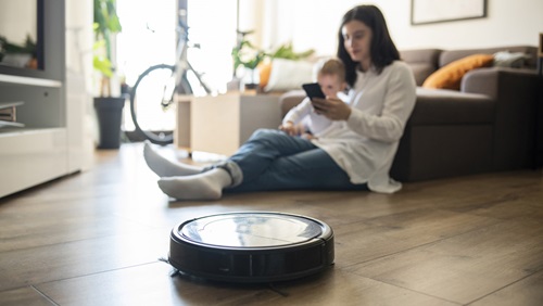 Een robotstofzuiger die door de woonkamer rijdt met vrouw met kind op schoot die naast de robotstofzuiger op de grond zit